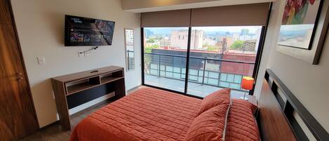 A bedroom with a large bed, a TV mounted on the wall, and a balcony with a city view.