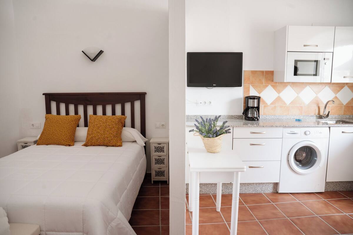 Un dormitorio con cama, mesitas de noche y una pequeña mesa con una planta. La habitación tiene una pared blanca y piso de baldosas.