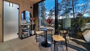 Ein modernes Zimmer mit einem großen Fenster, einem runden Tisch mit Stühlen, einer kleinen Bücherablage und einer Tür.