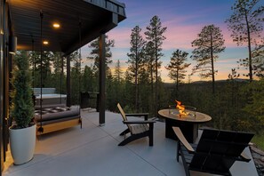 Une terrasse extérieure dotée d’un foyer, de sièges et offrant une vue sur une zone boisée.