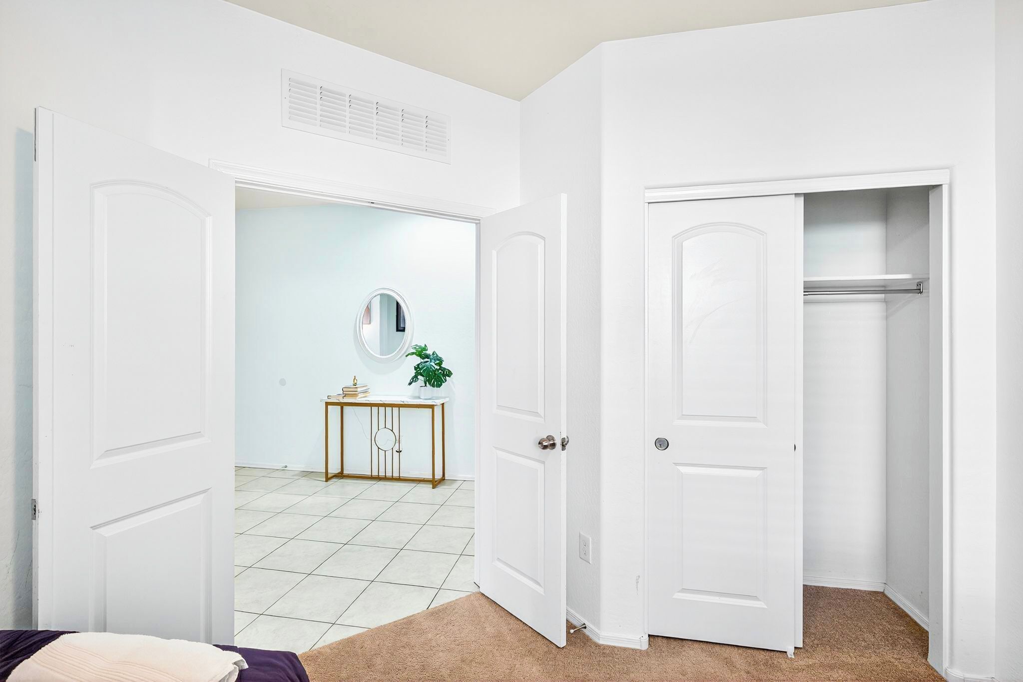 A room with white walls, a tiled floor, a bed with a beige blanket, a white door, and a small console table with a mirror and a plant.