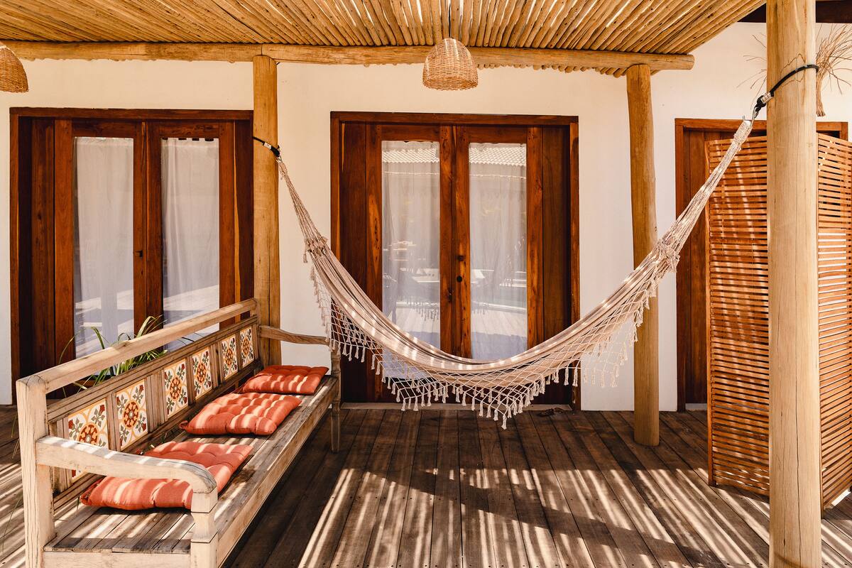 A hammock suspended between wooden posts in a room with a wooden bench and cushions, large windows with curtains, and a woven ceiling.
