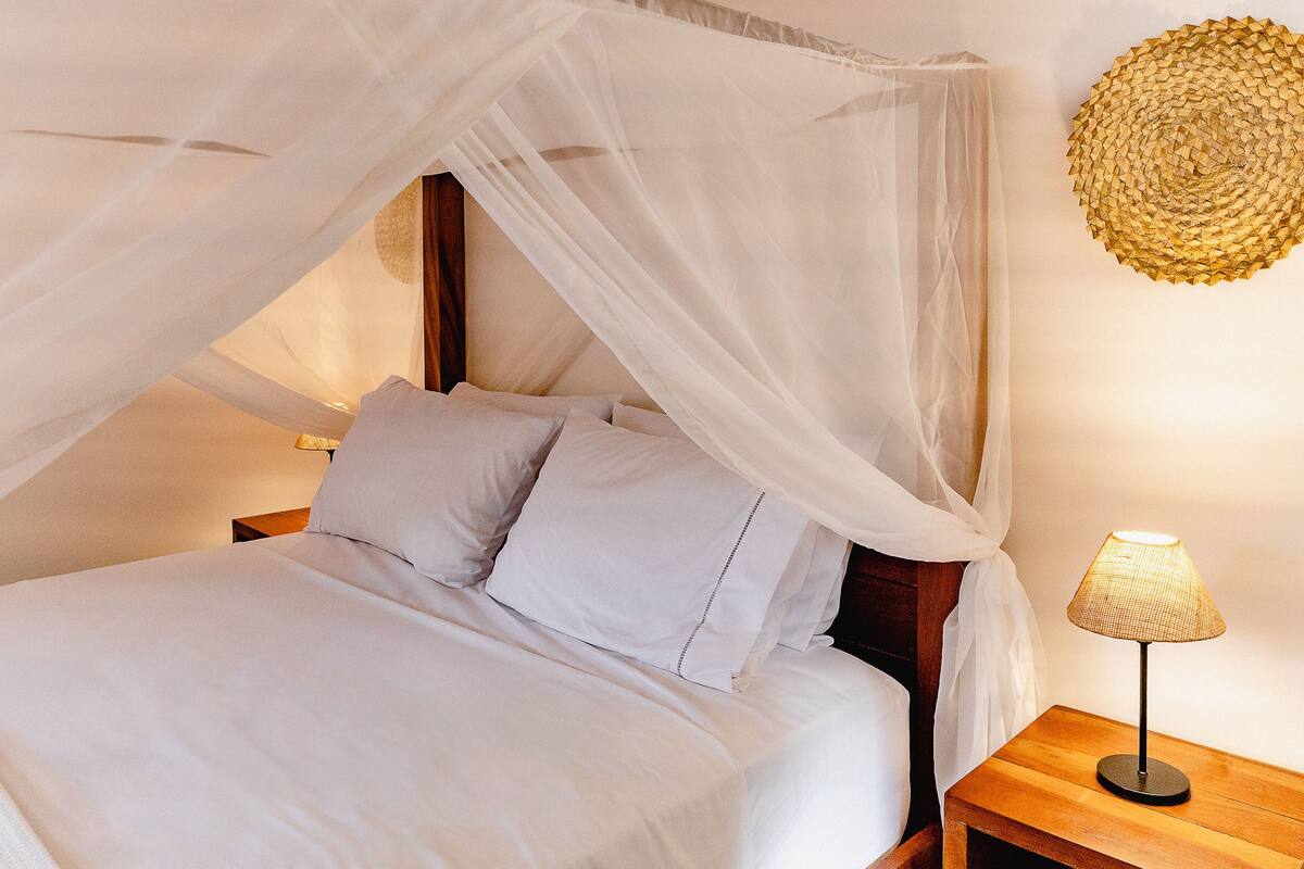 A bed with a canopy, a bedside lamp, and a woven wall art piece.