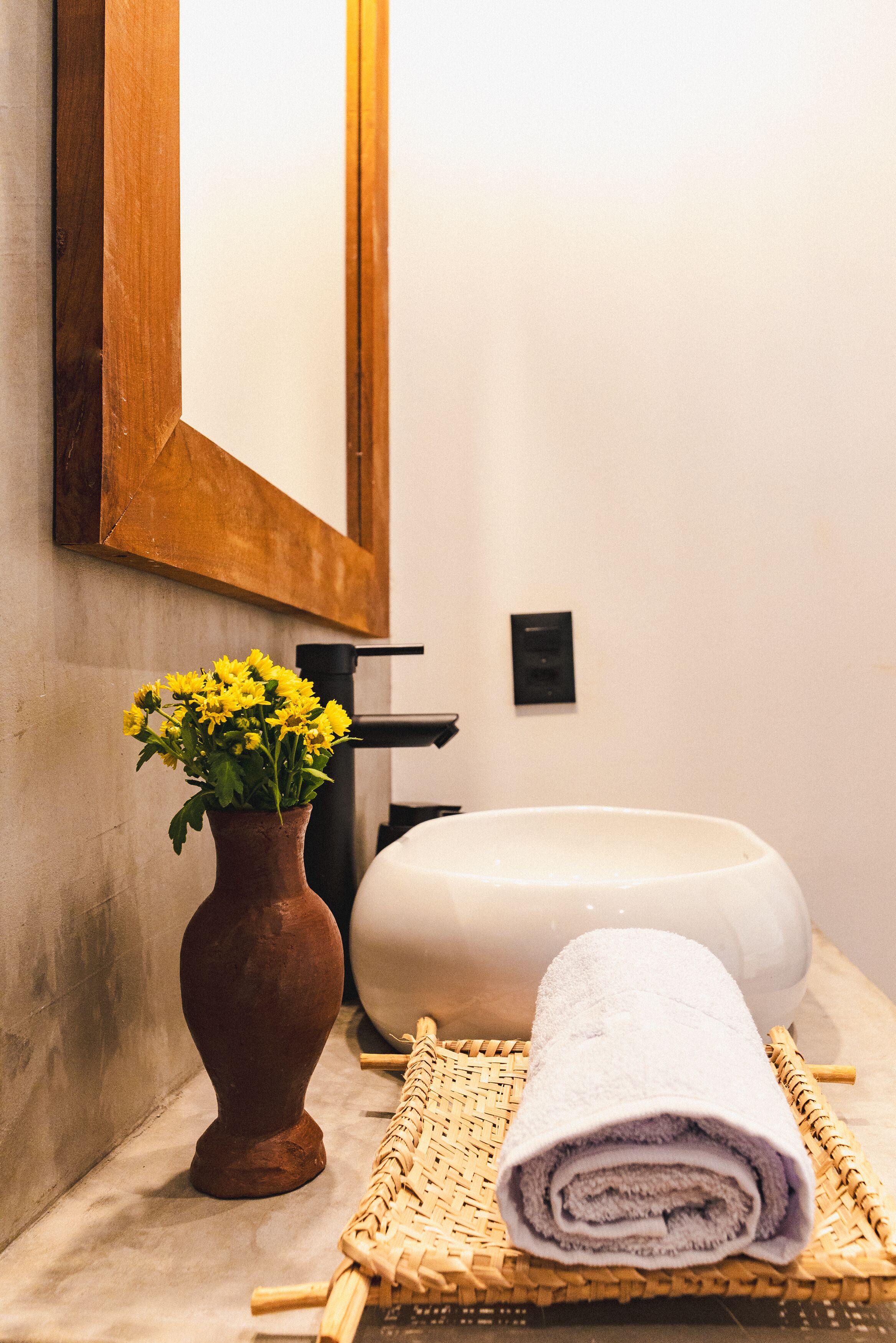 A bathroom with a white sink, a wooden-framed mirror, a vase with yellow flowers, and a rolled towel on a woven mat.