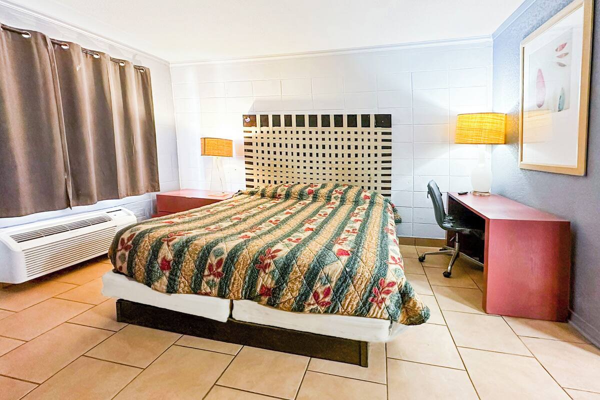 Quarto de hotel com cama, escrivaninha, cadeira, abajur e janela com cortinas.