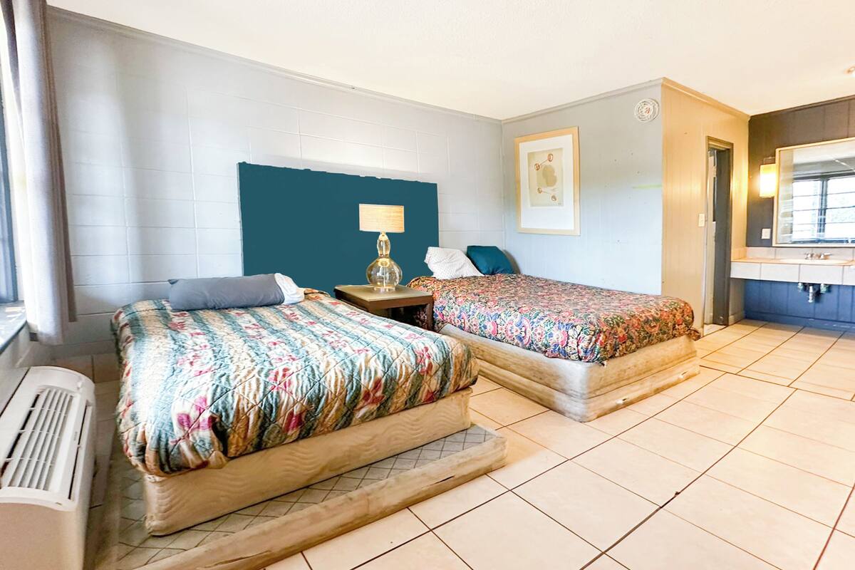 Quarto de hotel com duas camas, uma mesa de cabeceira, um abajur, um quadro na parede e um banheiro visível por uma porta aberta.