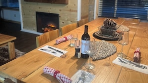 Una mesa de comedor de madera dispuesta con copas de vino, una botella y cubiertos, con una chimenea y un televisor al fondo.