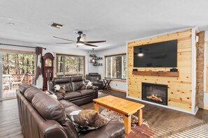 A modern living room with leather sofas, a wooden coffee table, a fireplace, and a large flat-screen TV mounted on a wooden wall.