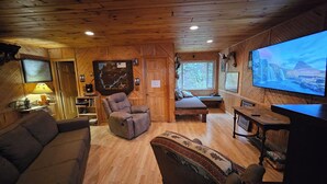 Interior acogedor de una cabaña con techo de madera, un sofá, un sillón reclinable, una mesita y un televisor que muestra una escena de naturaleza.