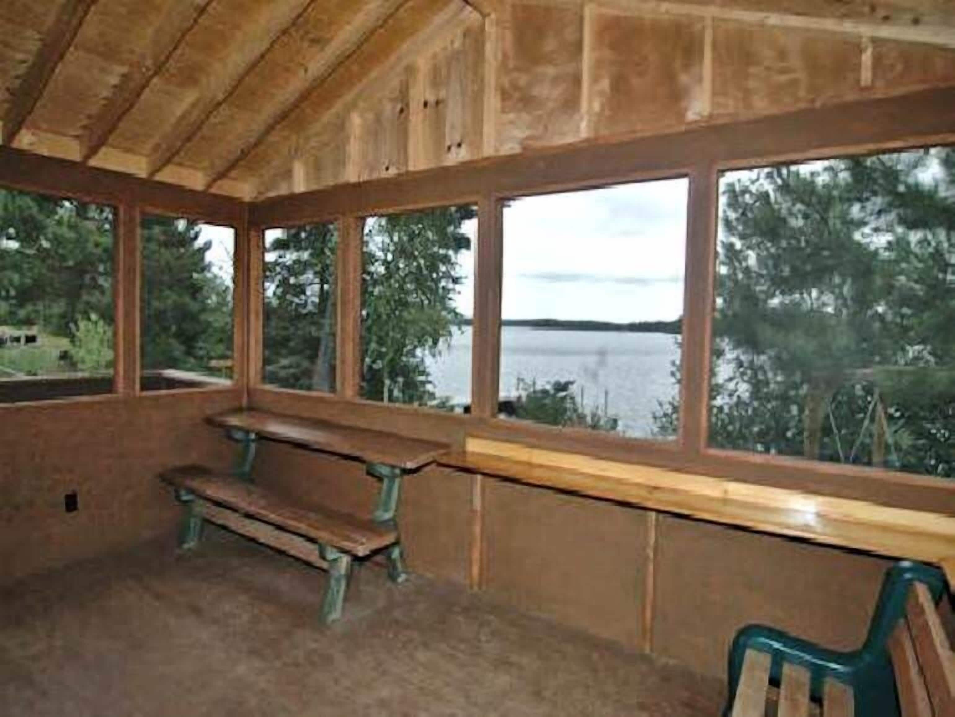 Une cabane en bois avec un banc et une table, de grandes fenêtres donnant sur un lac et des arbres.
