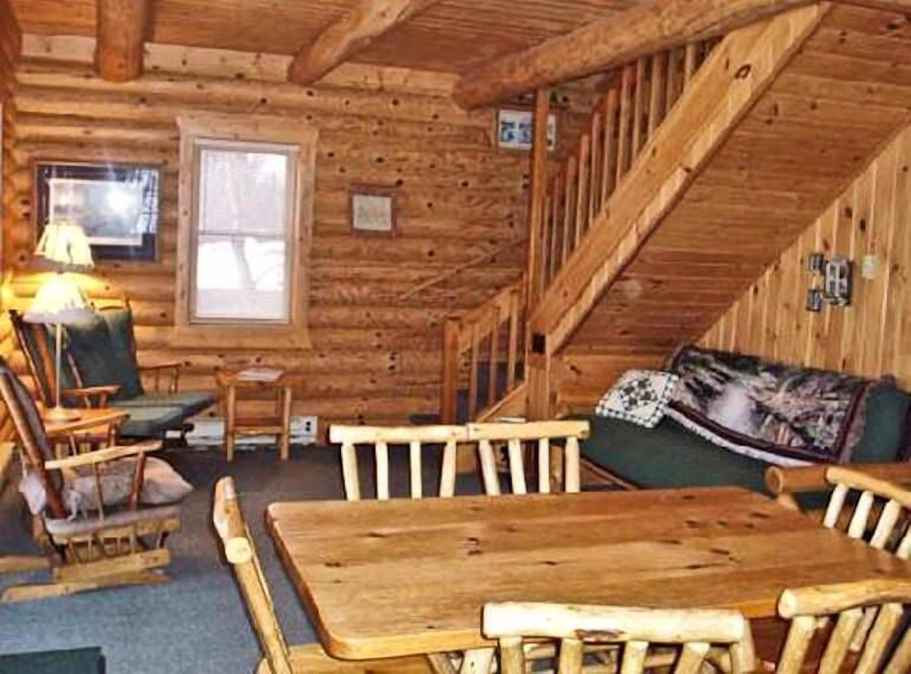 A log cabin interior with a wooden dining table, chairs, a sofa, and a staircase.