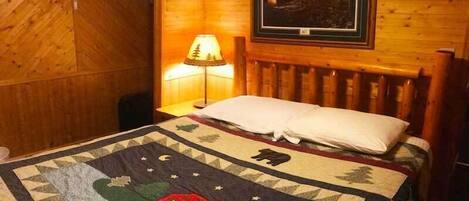 Una habitación acogedora con cabecera de madera, colcha con motivos campestres, lámpara e imagen enmarcada en la pared.
