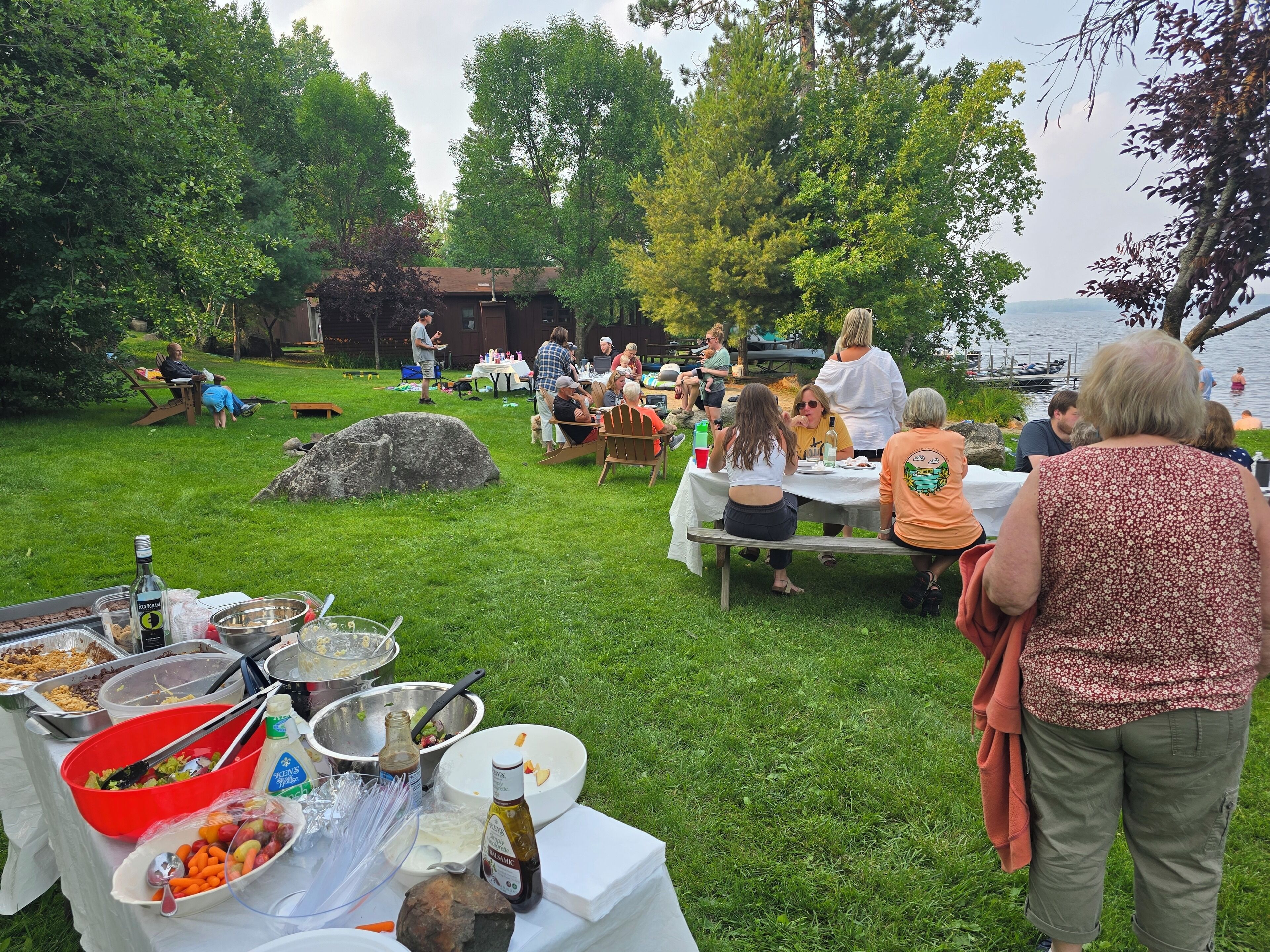 An outdoor dining area with tables set up on grass, surrounded by trees and a body of water.