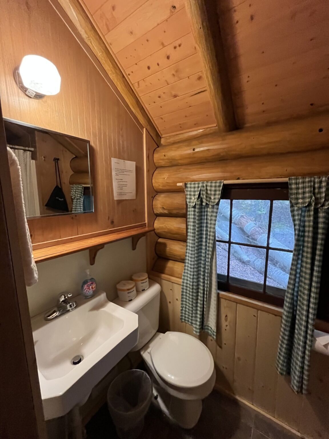 A wooden cabin bathroom with a sink, toilet, and a window with curtains.