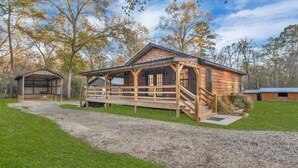 A wooden cabin with a porch and a covered area, surrounded by trees and a gravel path. - Cozy Country Cabin in the Woods (Dayton)