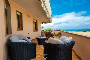 Un balcon agrémenté de meubles en osier, d’une table et de chaises, offrant une vue panoramique sur la mer et le paysage.