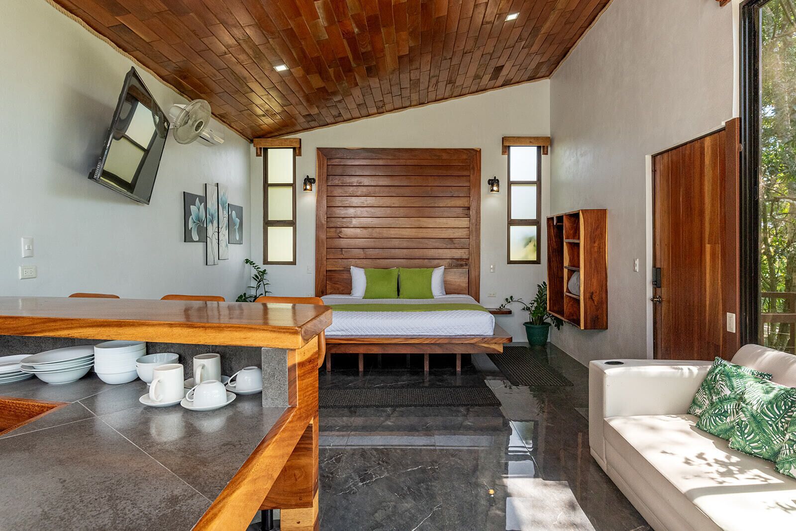 A modern room with a wooden ceiling, a bed with green pillows, a wooden bench, a dining table with white dishes, and a sofa with a green pillow.