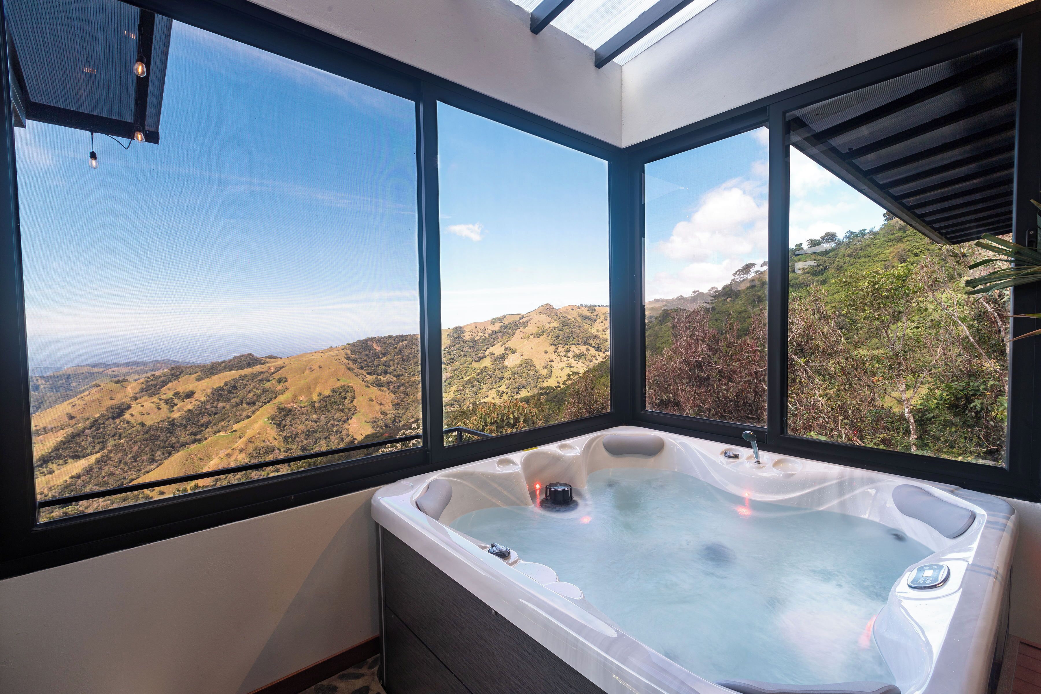 A hot tub with a panoramic view of a hilly landscape through large windows.