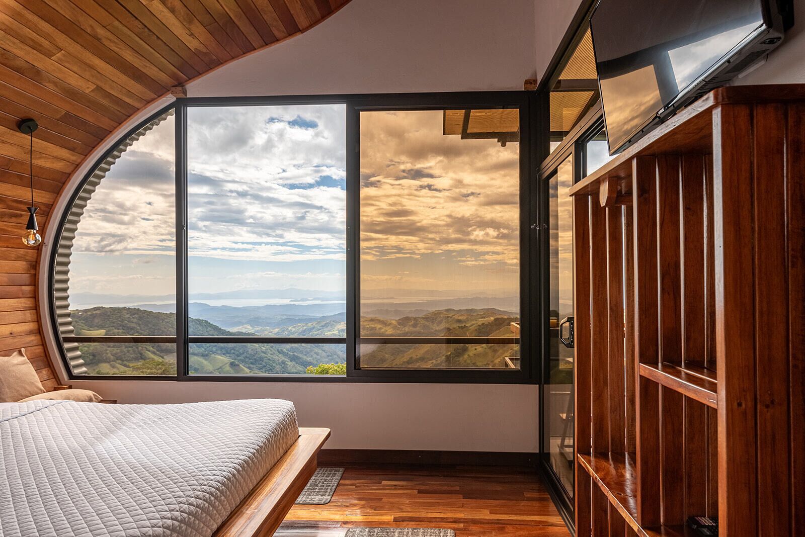 Una camera da letto con soffitto curvo, un'ampia finestra che offre una vista panoramica, un armadio in legno e un letto con copripiumino bianco.