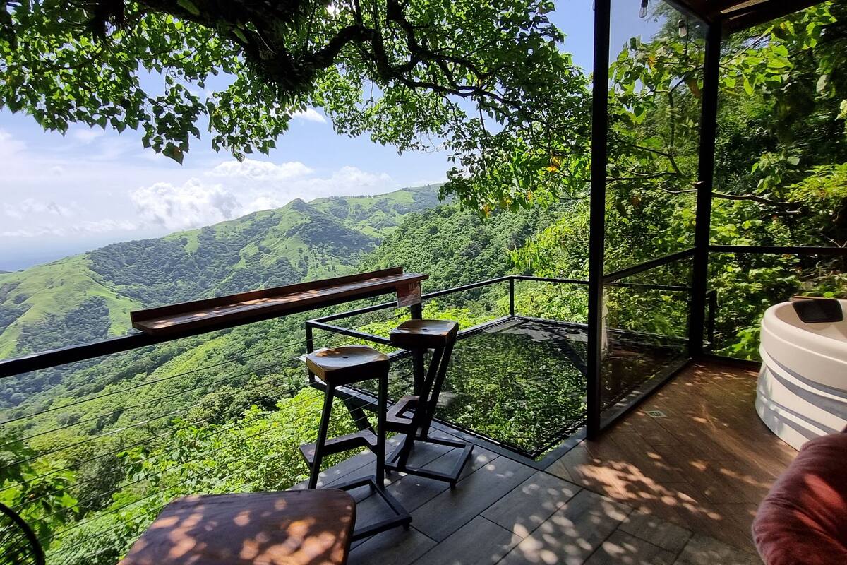 Un balcón con barandilla de vidrio que da a un exuberante valle verde y montañas distantes.