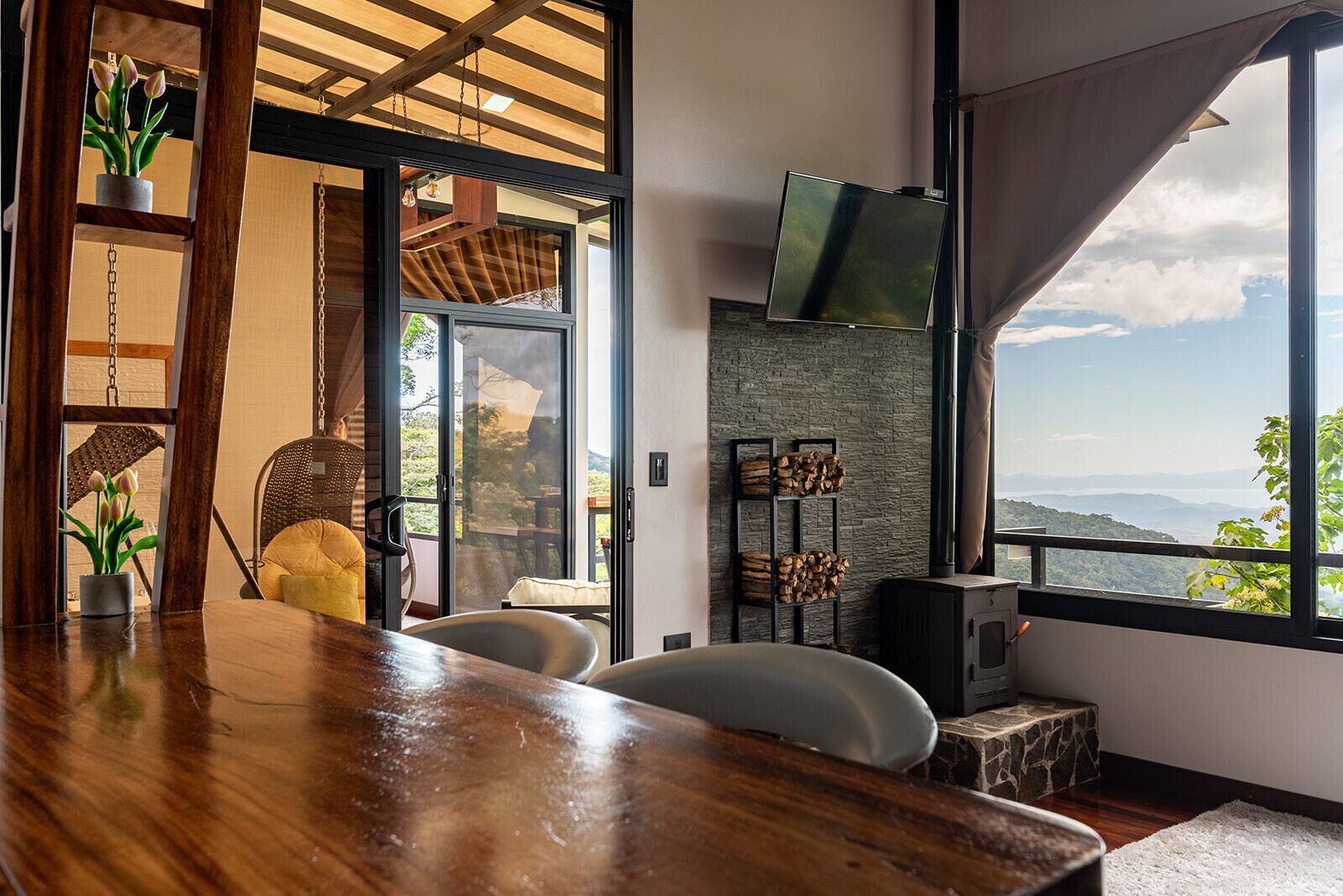 A modern living room with a wooden dining table, comfortable seating, a fireplace, and a large window offering a scenic view.