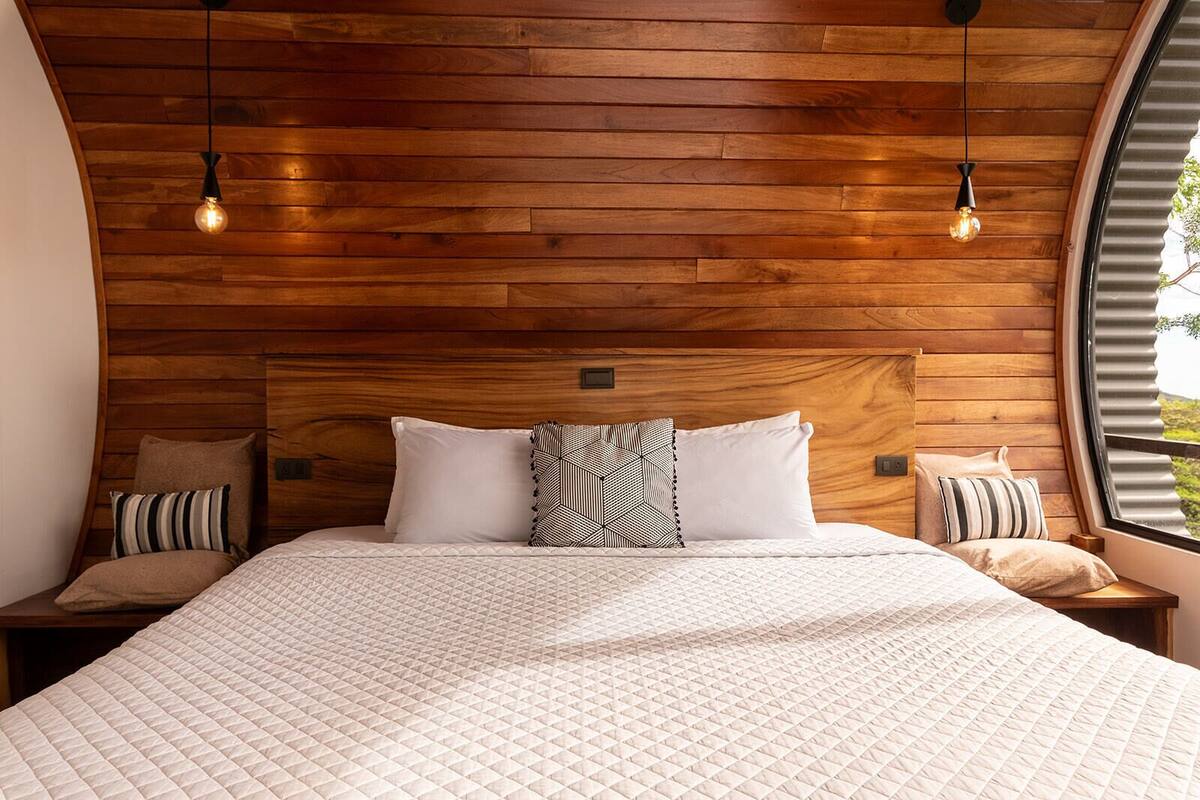 Un dormitorio moderno con techo de madera, una cama grande con edredón acolchado, dos mesitas de noche con lámparas y una ventana con vistas a zonas verdes.