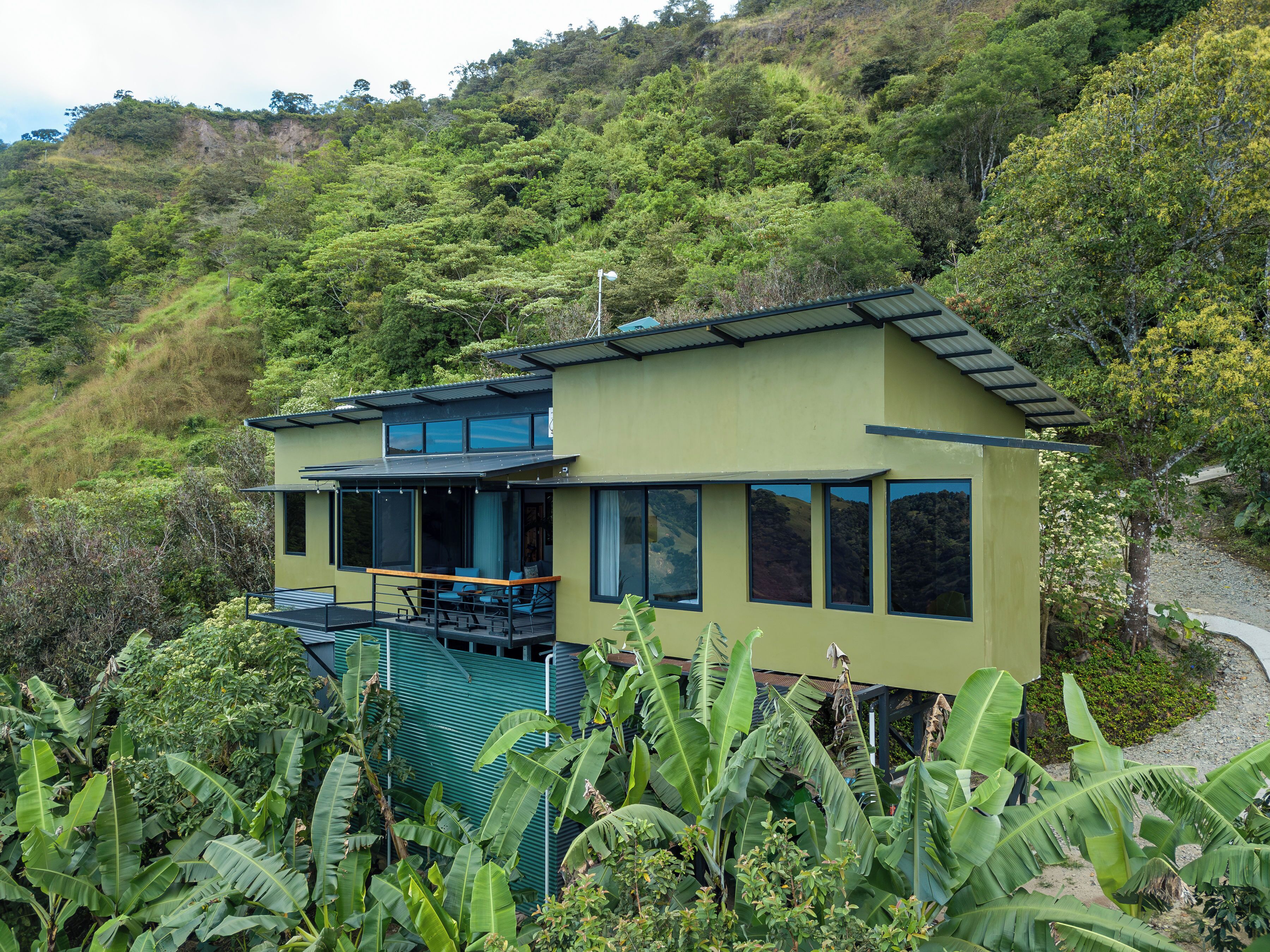 A modern two-story house with large windows and a balcony, surrounded by lush greenery and hills.