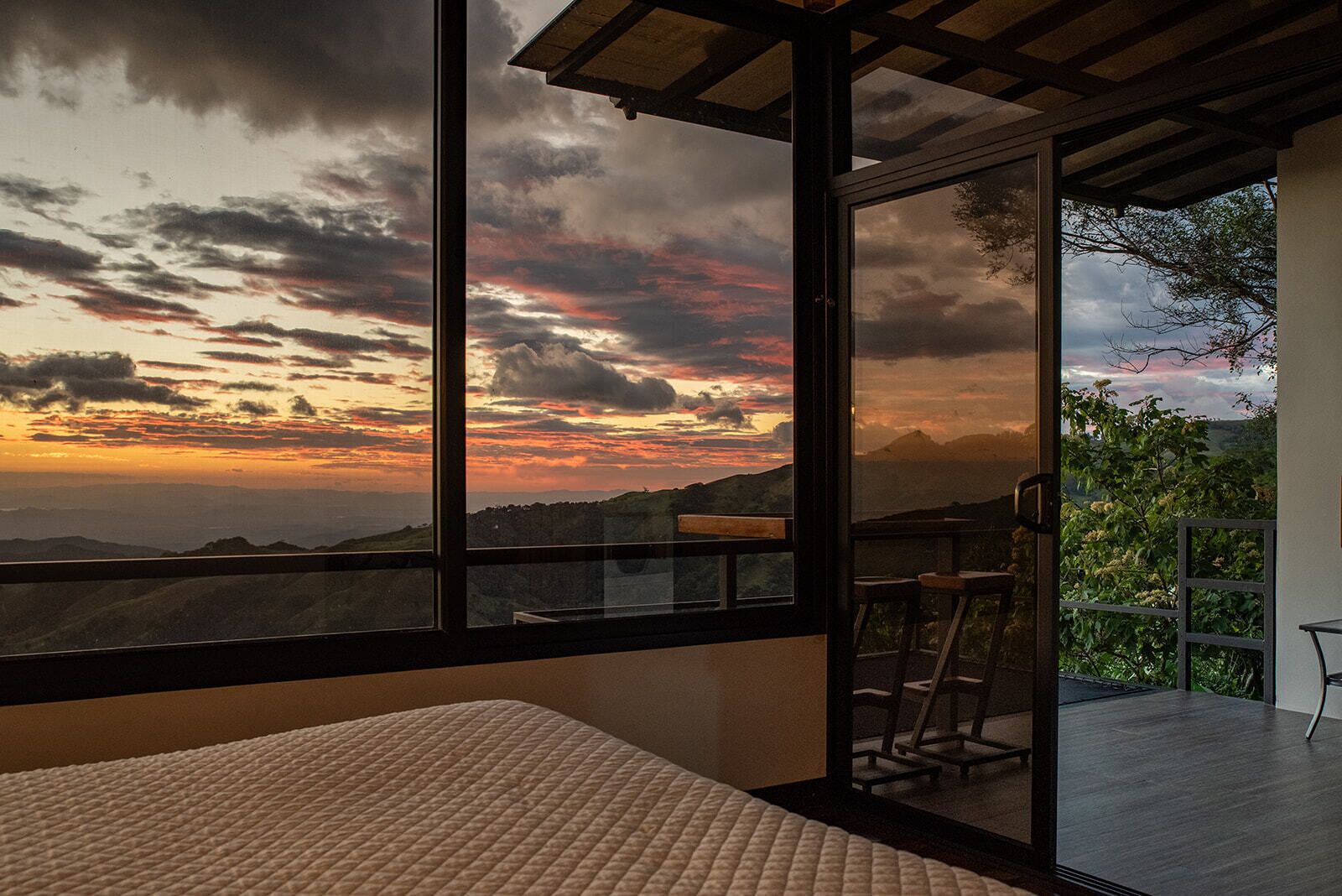 A room with a large window offering a view of a sunset over a hilly landscape.
