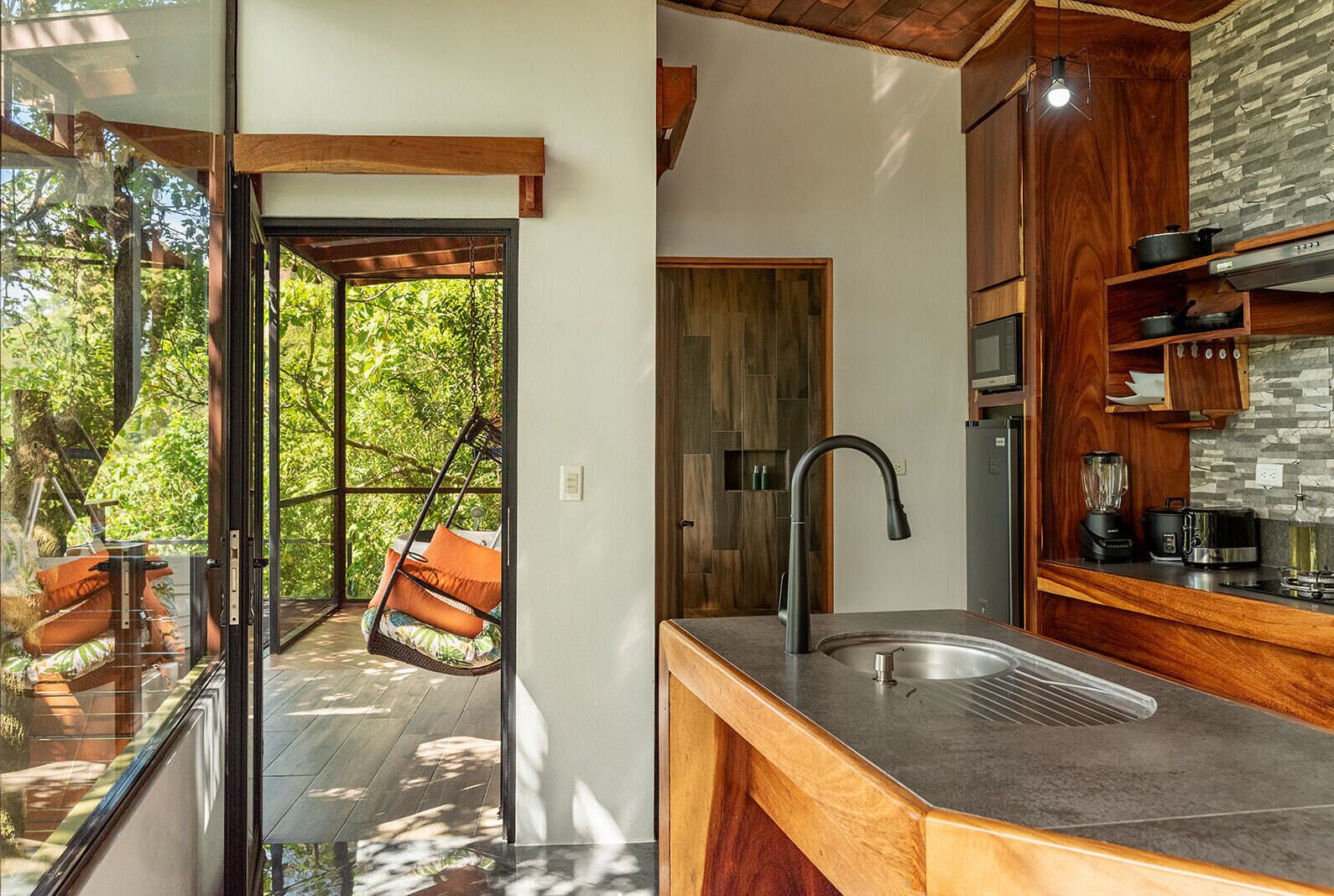 A modern kitchen with a large sink, wooden cabinets, and a hanging chair by a glass door leading to a balcony.