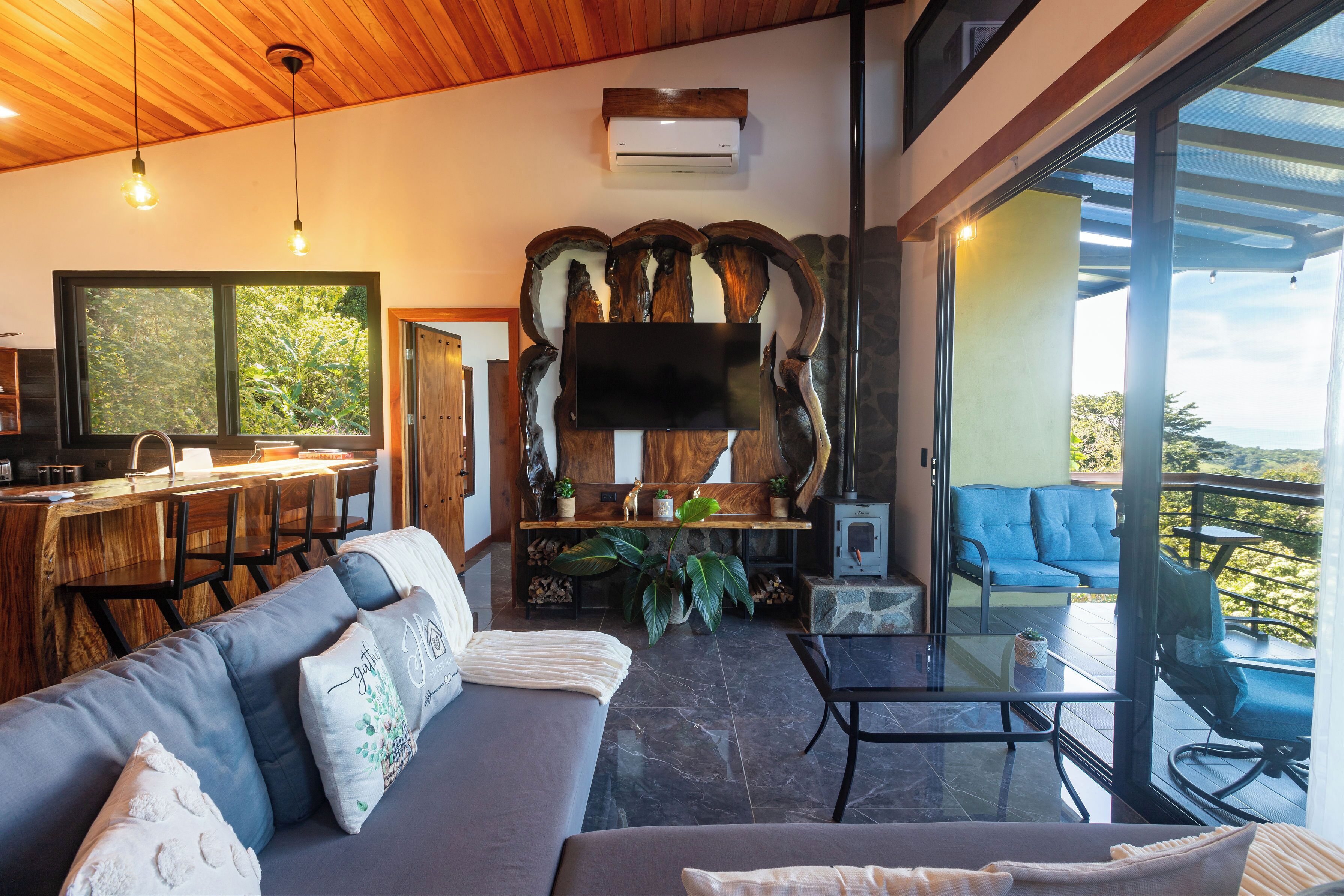 A modern living room with a sofa, a coffee table, a TV mounted on a wooden wall, a kitchen area with a bar, and a balcony with a blue chair.