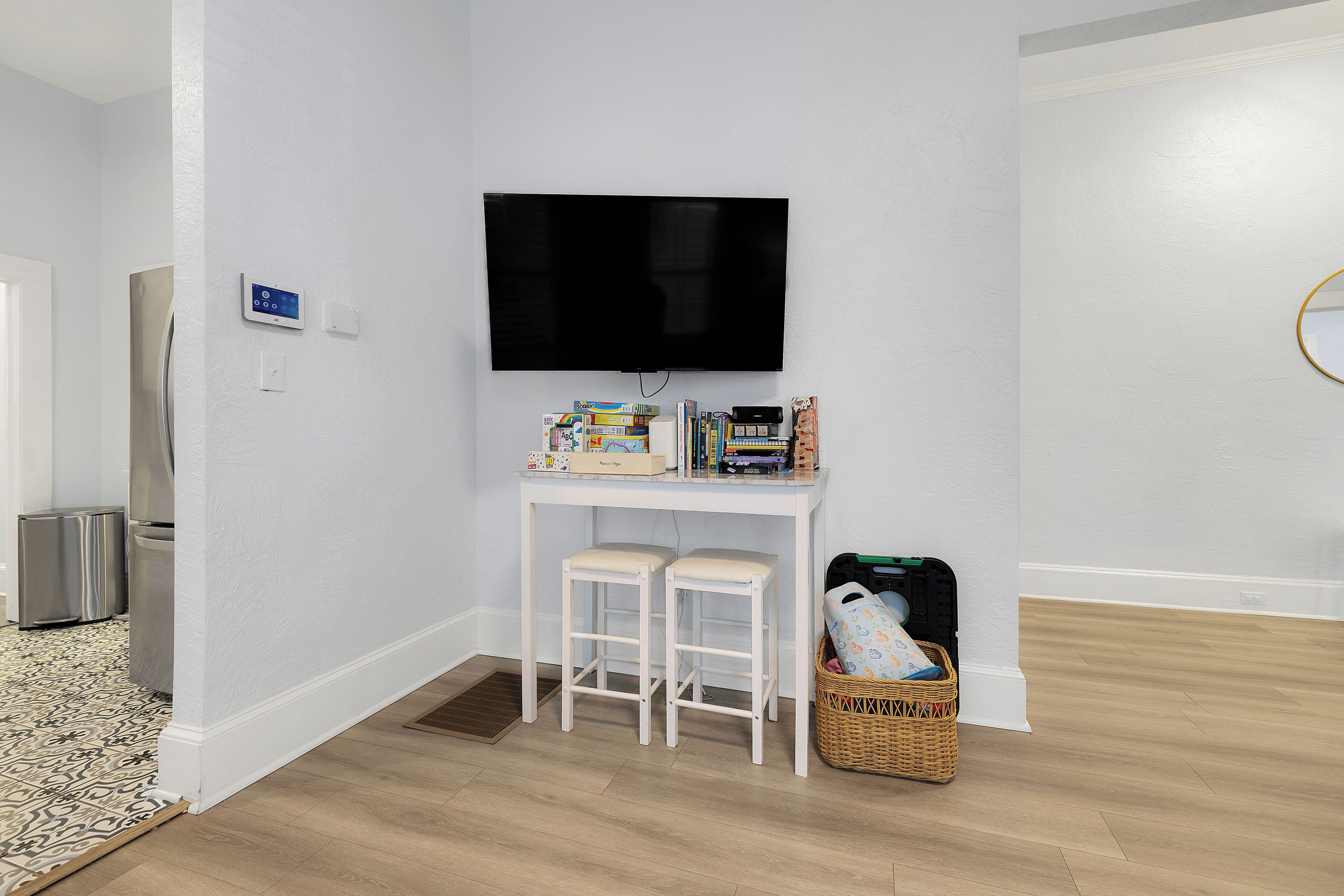 Una habitación con pared blanca, un televisor de pantalla plana sobre un escritorio blanco con dos taburetes, una cesta y varios objetos sobre el escritorio.