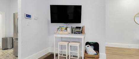 Una habitación con pared blanca, un televisor de pantalla plana sobre un escritorio blanco con dos taburetes, una cesta y varios objetos sobre el escritorio.