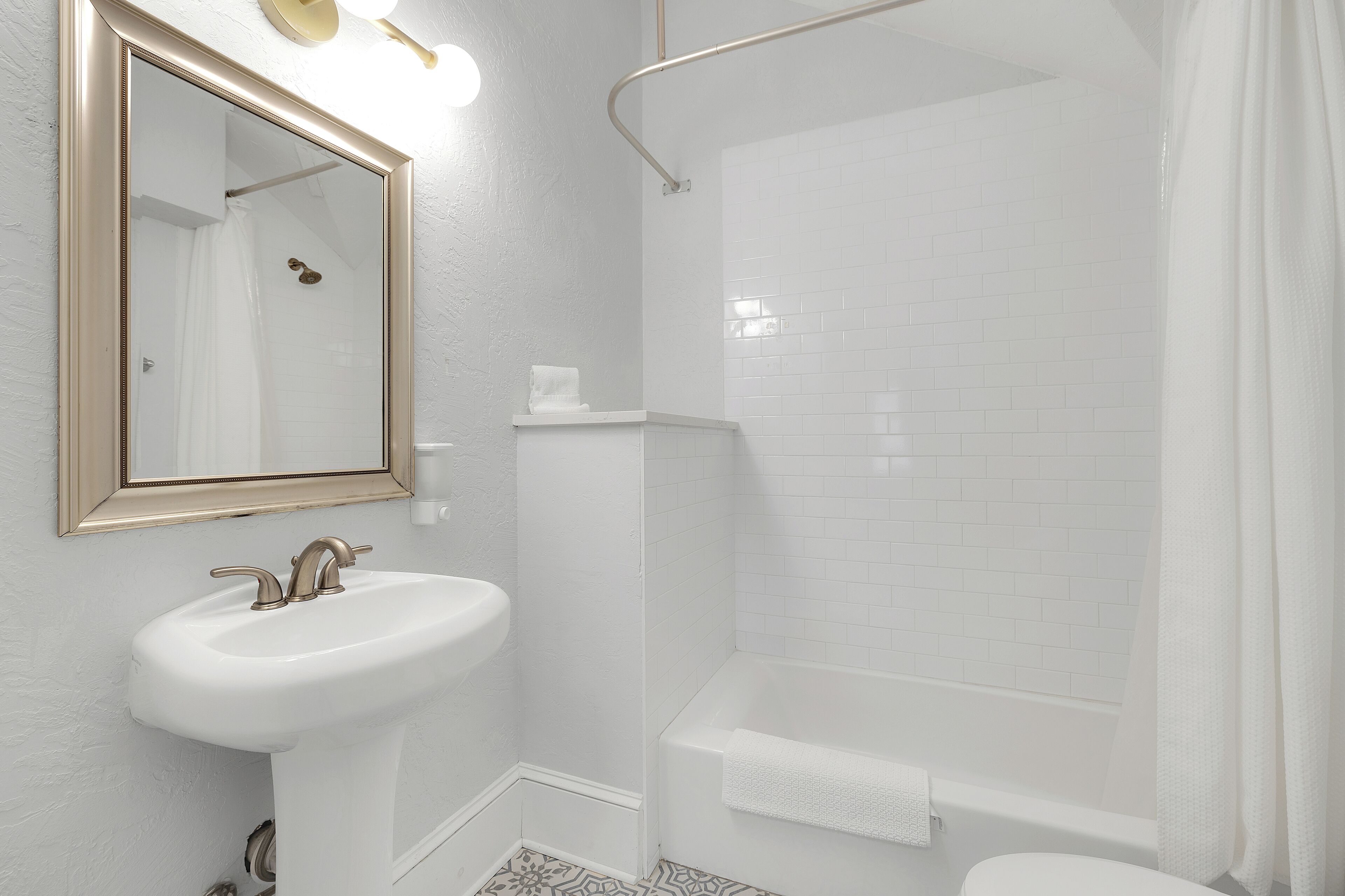 Ein Badezimmer mit einem weißen Waschbecken, einem Spiegel mit goldener Rahmung, einer Dusche mit weißem Vorhang und einer weißen Badewanne.