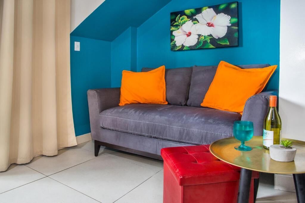 Salón con sofá gris, cojines naranja, un taburete rojo y una pequeña mesa redonda con una botella de vino y una copa.