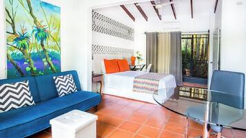 Habitación de hotel con sofá azul, cama con almohadas naranjas, escritorio y silla. En la pared hay un cuadro de una escena tropical, y el piso es de baldosas.