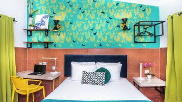 A hotel room with a bed, a desk with a laptop, a chair, a nightstand with a lamp, and a wall with a leaf pattern.