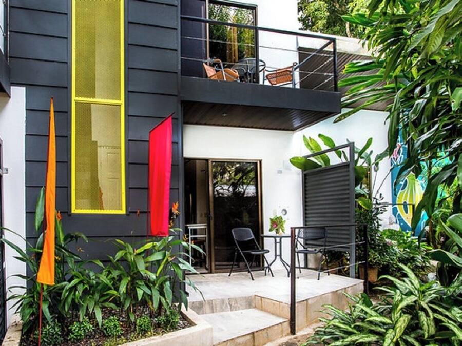 A modern two-story house with a balcony, outdoor seating, and lush greenery.