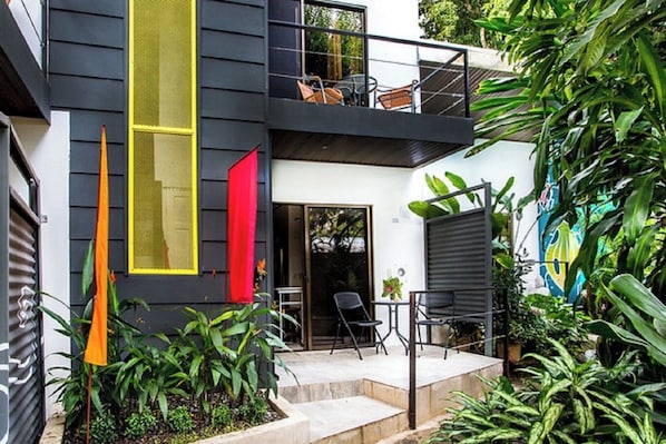 Una casa moderna de dos pisos con un balcón, mobiliario exterior y abundante vegetación.