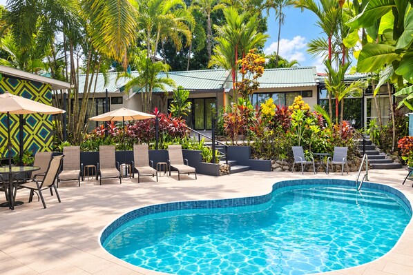 A swimming pool with lounge chairs and umbrellas, surrounded by tropical plants and a building with a green roof. - Tico Tico Villas Adult Only (Quepos)