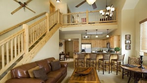 A spacious living area with a brown leather sofa, wooden coffee table, and a dining table with chairs. A kitchen with wooden cabinets and a bar area is visible in the background. - Cozy Suite with Gas Fireplace | Sauna + Spa (Park City)
