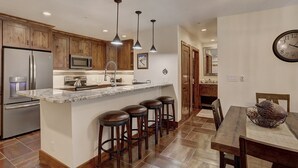 A kitchen with wooden cabinets, stainless steel appliances, a marble countertop, and a dining area with a wooden table and bar stools. - Premier Ski Condo w/ Slopeside Access (Breckenridge)