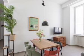 A modern living room with a wooden dining table, chairs, a potted plant, a TV, and a framed picture on the wall.