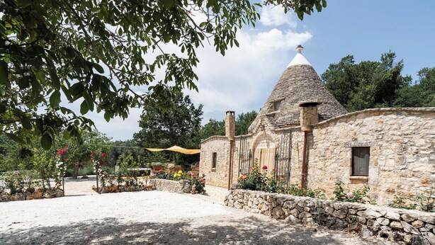 A stone building with a conical roof, surrounded by a gravel courtyard and flowering plants. - Trullo Fragno by Wonderful Italy (Noci)