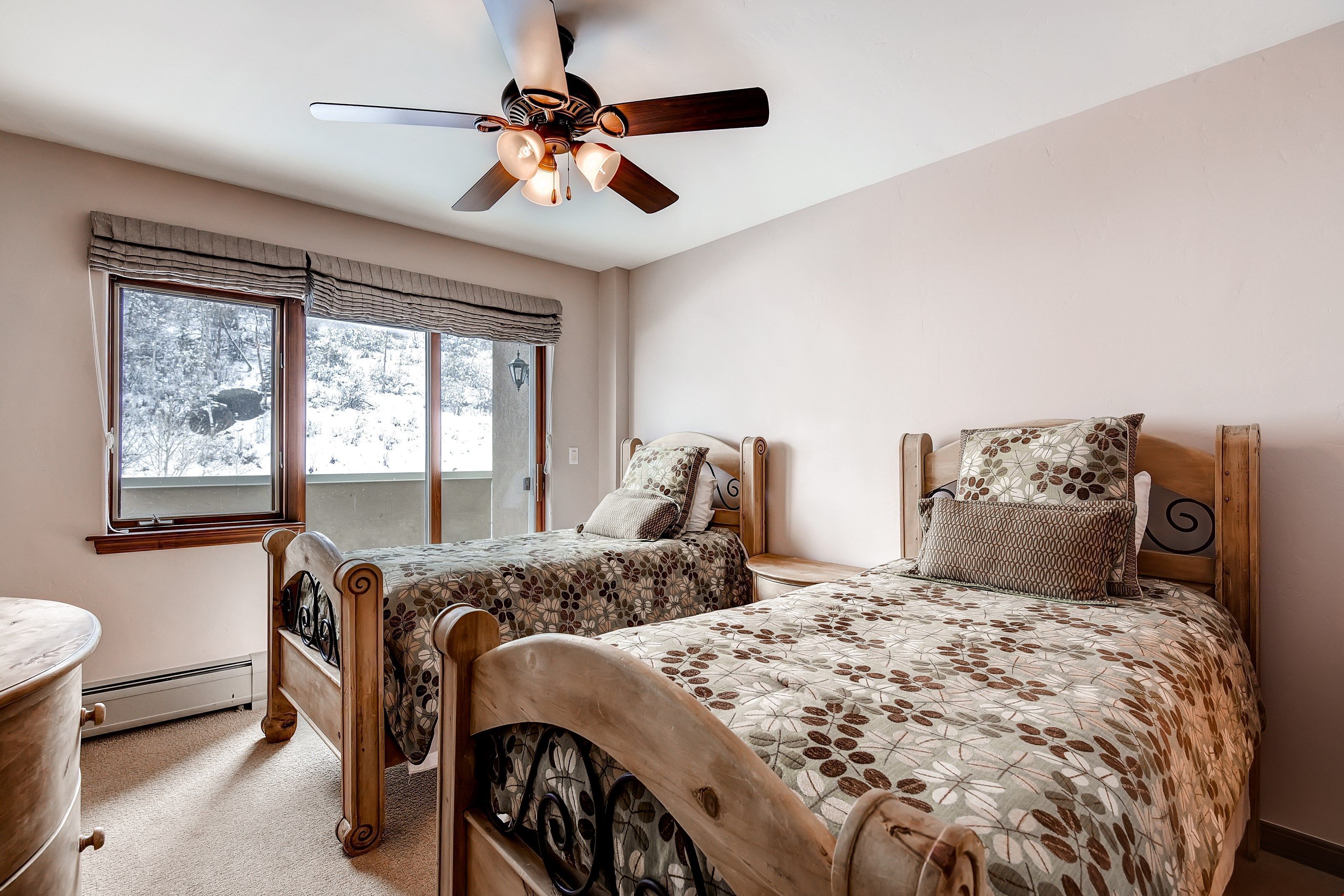 A bedroom with two wooden beds, a ceiling fan, and a large window with a view of snow-covered trees.