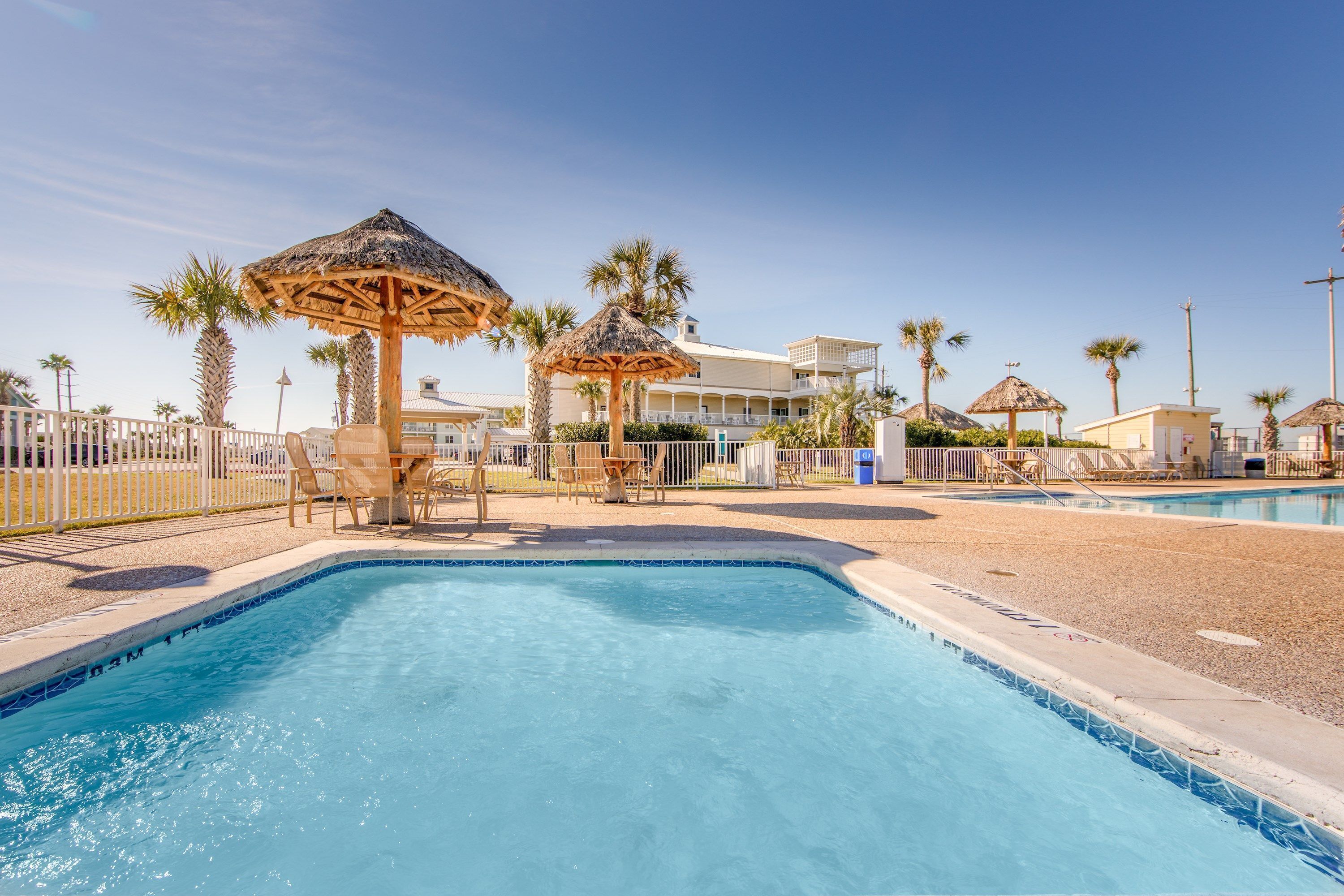 A swimming pool with lounge chairs and thatched umbrellas, surrounded by palm trees and a building with balconies.