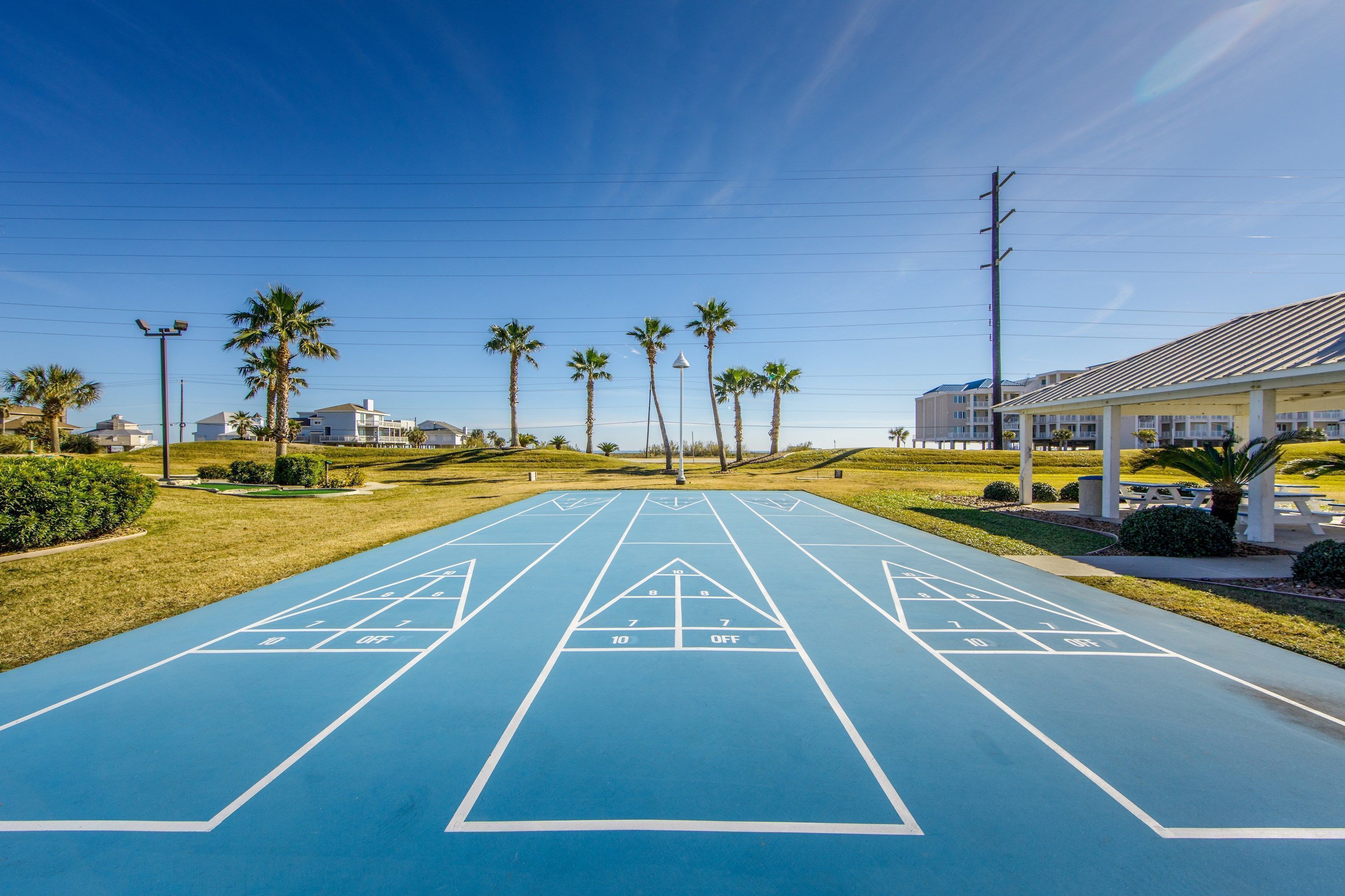 A blue shuffleboard court with numbers and a 'OFF' sign, surrounded by palm trees and a building with a sloped roof.