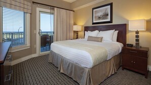 Une chambre d’hôtel avec un grand lit, deux tables de chevet avec des lampes, un bureau, une chaise, un balcon avec vue et un tableau encadré au mur.