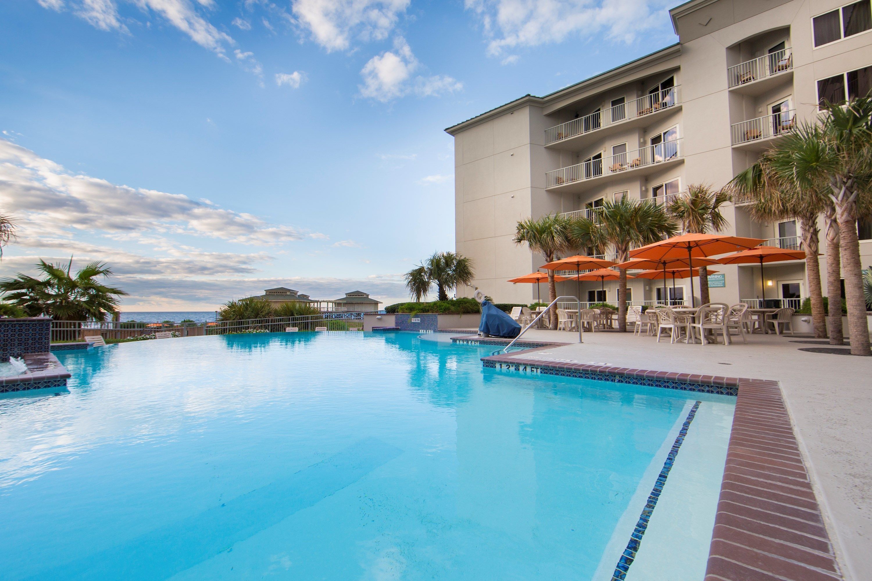 A swimming pool with a view of the ocean, surrounded by palm trees and outdoor seating.