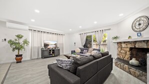 Uma sala de estar moderna com um sofá modular cinza, uma TV de tela plana, uma lareira de pedra e uma janela grande com cortinas.