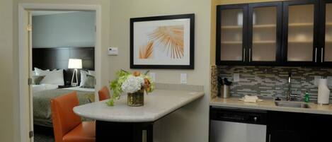 A hotel room with a bed, a desk with a chair, a small kitchen area with cabinets and a dishwasher, and a framed picture on the wall.